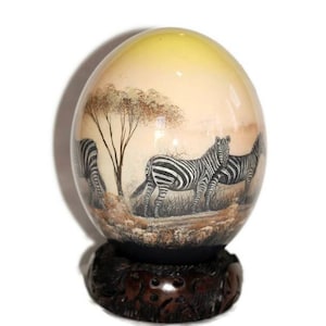 May include: An egg-shaped decorative object with a painted scene of zebras in a savanna landscape. The egg is mounted on a dark brown, carved wooden base. The artwork features a tree and a warm-toned sky.