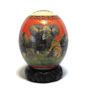 May include: A hand-painted ostrich egg with a red background and a scene of African animals, including an elephant, lion, and leopard. The egg is displayed on a wooden stand.
