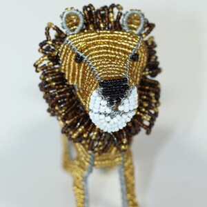 May include: A beaded lion figurine with a gold and brown mane, black eyes, and a white muzzle. The lion is standing on a white surface.