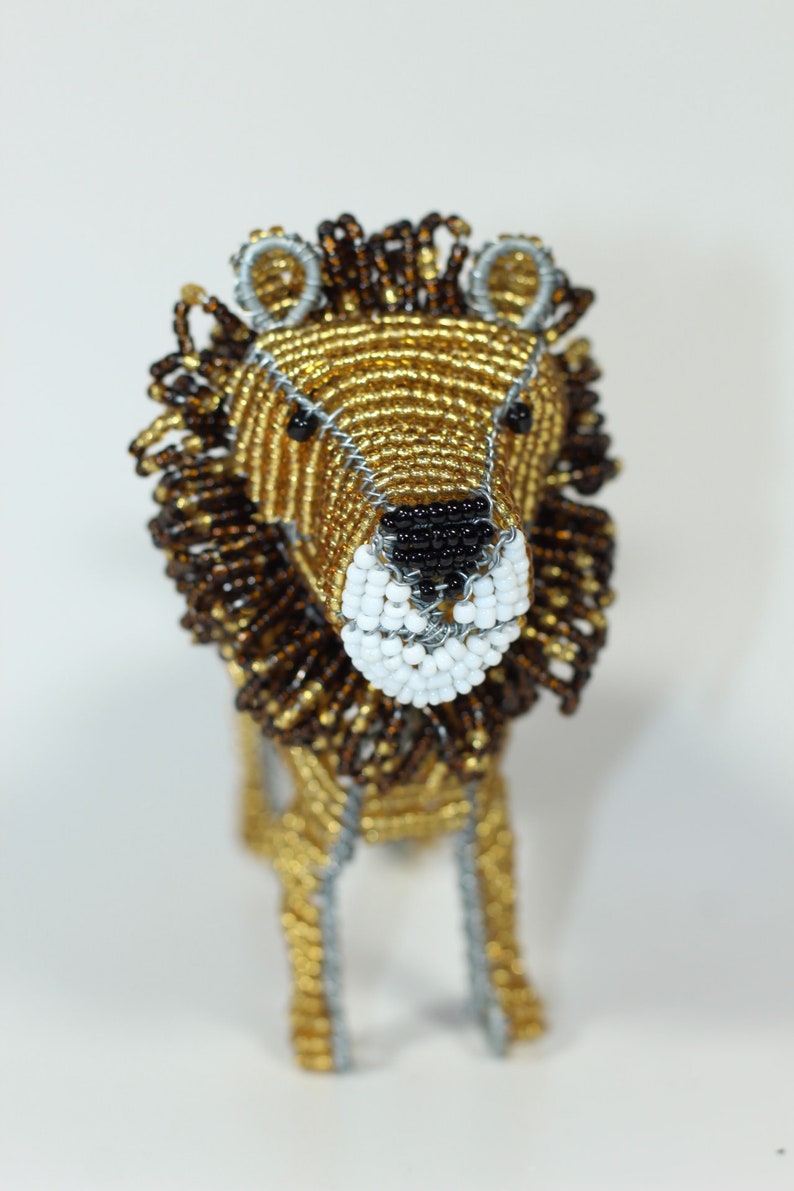 May include: A beaded lion figurine with a gold and brown mane, black eyes, and a white muzzle. The lion is standing on a white surface.
