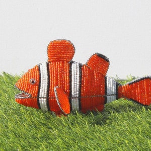 Puede incluir: Una figurita de pez payaso naranja y blanco hecha de cuentas. El pez está hecho de cuentas pequeñas y alambre y tiene una forma realista.