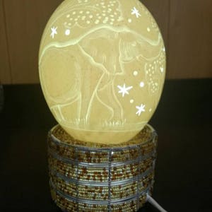 Op de afbeelding: Een witte struisvogel-eierlamp met een gegraveerd olifantenmotief op het oppervlak. De lamp is verlicht en heeft een kralenbasis.