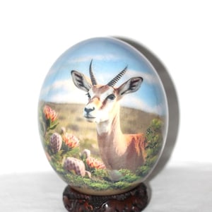 May include: An oval decorative egg featuring a painted gazelle in a natural landscape with flowers and a blue sky. The egg is mounted on a dark brown wooden base, creating a unique art piece.
