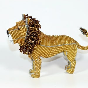 Puede incluir: Una figurita de león de cuentas con cuerpo dorado, melena marrón y detalles de alambre plateado. El león está de pie sobre sus cuatro patas con la cola enroscada.