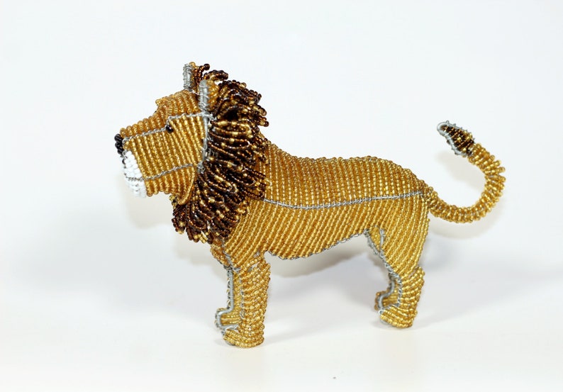 A beaded lion figurine with a gold body, brown mane, and silver wire accents. The lion is standing on all four legs with its tail curled up behind it. Handmade in South Africa by Job Guwhe