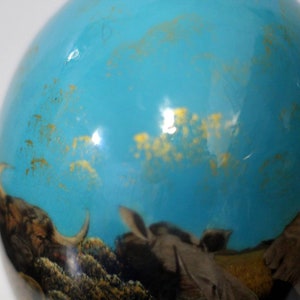 May include: A blue glass ornament with a gold flecked design. The ornament features a scene of a buffalo and other animals in a grassy field.