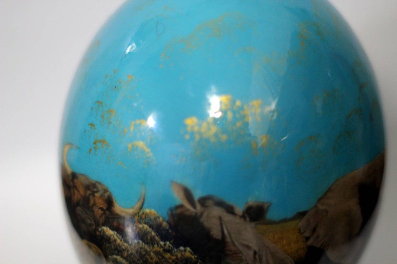 May include: A blue glass ornament with a gold flecked design. The ornament features a scene of a buffalo and other animals in a grassy field.