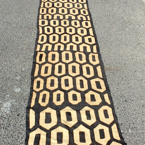 Puede incluir: Una alfombra larga, tejida en negro y beige con un patrón geométrico de hexágonos repetidos. La alfombra está hecha de una tela gruesa y texturizada y tiene flecos en los extremos.