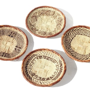 May include: Four woven baskets with brown rims and intricate black and white patterns. The baskets are round and shallow, and they are arranged in a circle on a white background.