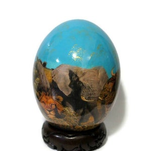 May include: A hand-painted ostrich egg with a blue background and a scene of African animals, including elephants, lions, and a rhinoceros. The egg is displayed on a dark wooden stand.