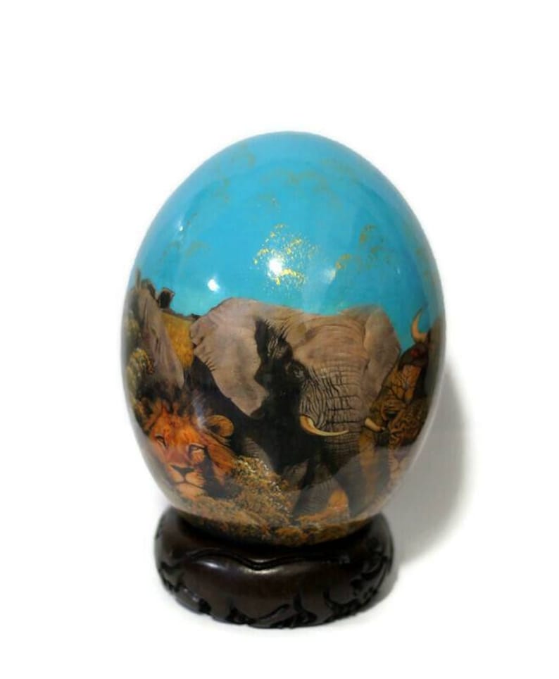 May include: A hand-painted ostrich egg with a blue background and a scene of African animals, including elephants, lions, and a rhinoceros. The egg is displayed on a dark wooden stand.