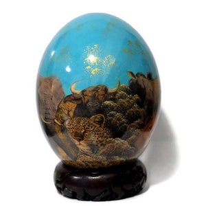 May include: A hand-painted ostrich egg with a blue background and a scene of African animals, including an elephant, a leopard, and a buffalo. The egg is displayed on a wooden stand.