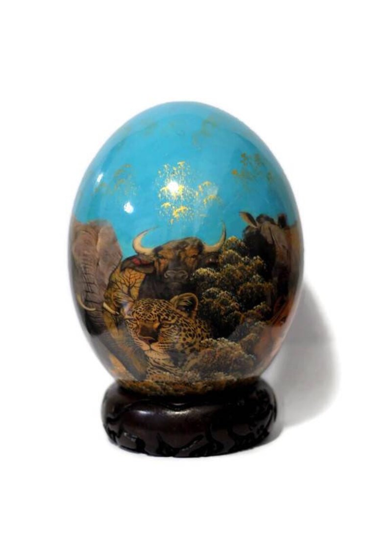 May include: A hand-painted ostrich egg with a blue background and a scene of African animals, including an elephant, a leopard, and a buffalo. The egg is displayed on a wooden stand.