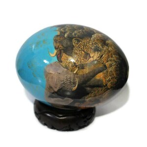 May include: A hand-painted, blue and brown egg-shaped ornament with a scene of a leopard, elephant, and rhino in a jungle setting. The ornament is on a dark wood stand.