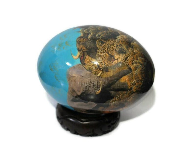 May include: A hand-painted, blue and brown egg-shaped ornament with a scene of a leopard, elephant, and rhino in a jungle setting. The ornament is on a dark wood stand.