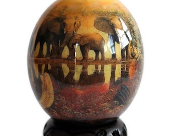 May include: An oval-shaped decorative object featuring a detailed illustration of elephants standing near a water source at sunset, with warm orange and brown tones. The object is mounted on a dark, circular, carved wooden base with intricate patterns.