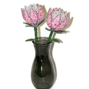 May include: Two pink and white beaded protea flowers in a dark grey vase. The flowers are made of beads and wire and have a realistic appearance.