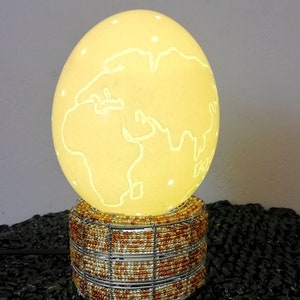 Op de afbeelding: Een witte struisvogel-eierlamp met een wereldkaartontwerp dat in het oppervlak is gegraveerd. De lamp staat op een kralenbasis met een draadframe.