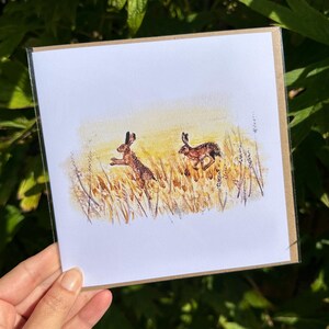 May include: A greeting card featuring a watercolour illustration of two brown rabbits hopping through a field of tall yellow grass.