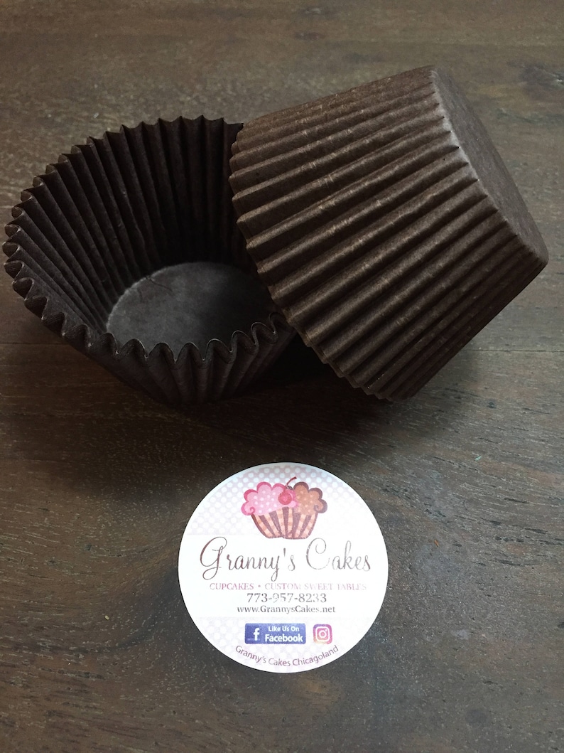 May include: Two brown paper cupcake liners, one partially inside the other, on a wooden surface. A white sticker with a pink cupcake graphic and the text "Granny's Cakes" is below the liners.