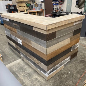 May include: A corner bar counter with a light wood top and a rustic wood front. The front is made of reclaimed wood planks in various shades of brown, gray, and white.