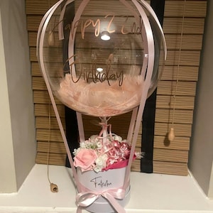 May include: A clear balloon shaped like a hot air balloon with pink feathers inside, featuring the text Happy 22nd Birthday in rose gold. It sits atop a white cylindrical box labelled Fateha, decorated with pink and white artificial roses and ribbons.