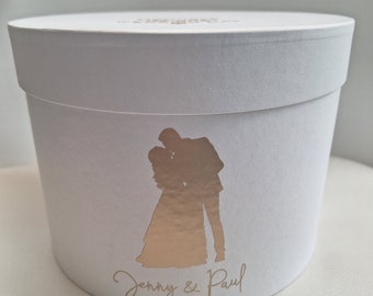 May include: White round box with a gold silhouette of a couple embracing. The box is decorated with the text "Jenny & Paul" and the date "20/08/2023".
