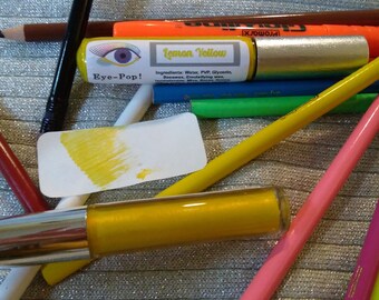 May include: A close-up of a yellow liquid eye shadow in a silver tube with the label "Lemon Yellow" and "Eye-Pop!"  The tube is surrounded by various colored pencils and markers.