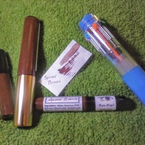 May include: An assortment of brown eye makeup products and a blue multi-color pen are arranged on a green surface. The makeup includes a lip gloss, a tube with a gold base, and a dual-ended eye pencil labeled "Spiced Brown" and "Eye-Pop!"