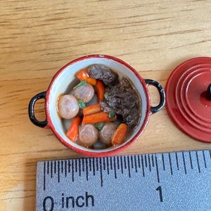 May include: A miniature red round pot with black handles containing a simulated stew of brown meat chunks, orange carrot slices, and round beige potatoes. A matching red lid sits beside the pot next to a ruler showing a diameter of 1 inch.