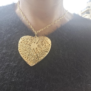 May include: Gold-toned heart pendant necklace. The pendant features an intricate, openwork design. The necklace has a delicate chain. The pendant is large and eye-catching, making it a statement piece. The necklace is worn against a dark background.