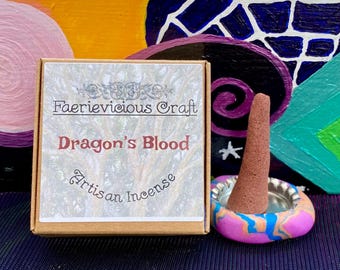Może przedstawiać: Małe pudełko z napisem "Faerievicious Craft Dragon's Blood Artisan Incense" na przodzie. Czerwony stożek kadzidła znajduje się na kolorowym ceramicznym stojaku.