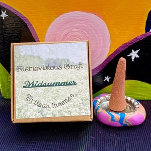 Op de afbeelding: Een bruine doos met de tekst "Faerievicious Craft Midsummer Artisan Incense". Een roze, blauwe en turquoise swirl keramische wierookhouder met een bruine wierookkegel.
