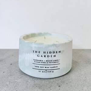 May include: A round, light green and white candle with three wicks. The candle has the text "THE HIDDEN GARDEN" and lists the scents: Gardenia, Beach Linen, Sugar Pine & Patchouli. It is a 12 oz (340 g) soy wax candle.