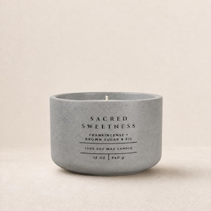 May include: A gray cylindrical candle in a textured container with a single wick. The label reads SACRED SWEETNESS, FRANKINCENSE + BROWN SUGAR & FIG, 100% SOY WAX CANDLE, 12 OZ | 340 g. The candle has a smooth, matte finish and a centered white wick.