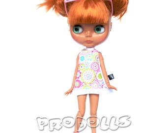 May include: A doll with orange hair, green eyes, and pink glasses wearing a white dress with a colorful floral pattern. The doll is wearing pink shoes and has pigtails.