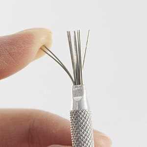 Könnte beinhalten: Ein silbernes Werkzeug aus Metall mit mehreren dünnen, spitzen Drähten, die sich vom Ende erstrecken. Die Drähte werden zum Modellieren und Detaillieren von Ton verwendet.