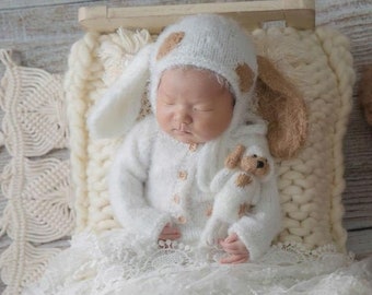 May include: A newborn baby wearing a white knitted bunny costume with brown accents, lying on a white knitted bed with a white lace trim. A brown poodle dog is sitting on the wooden floor next to the bed.