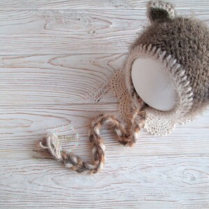 Peut inclure: Un bonnet b&eacute;b&eacute; en crochet marron et cr&egrave;me avec des oreilles en fourrure et une longue cravate en crochet. Le bonnet est pos&eacute; sur une surface en bois blanc.