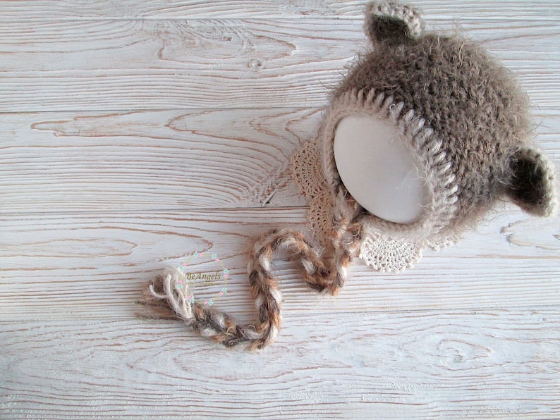 Peut inclure: Un bonnet b&eacute;b&eacute; en crochet marron et cr&egrave;me avec des oreilles en fourrure et une longue cravate en crochet. Le bonnet est pos&eacute; sur une surface en bois blanc.
