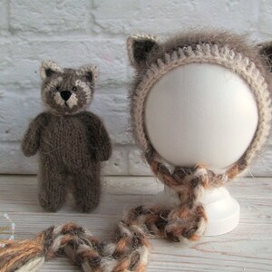 Peut inclure: Un bonnet en tricot marron en forme de raton laveur avec des oreilles duveteuses et une bordure blanche en crochet. Le bonnet est sur une t&ecirc;te de mannequin blanche et a une queue en laine marron et blanche.