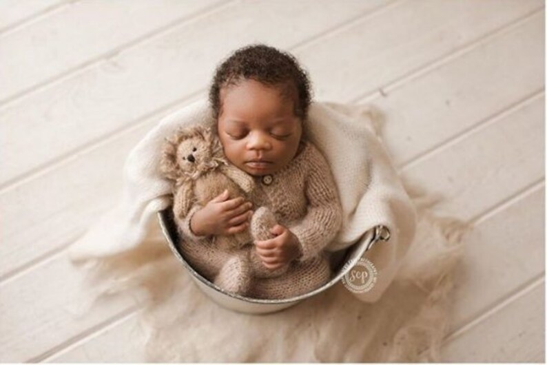 Puede incluir: Un beb&eacute; reci&eacute;n nacido con un mono de punto marr&oacute;n est&aacute; durmiendo en un cubo blanco. El beb&eacute; est&aacute; sosteniendo un animal de peluche marr&oacute;n. El beb&eacute; est&aacute; rodeado de una manta blanca.