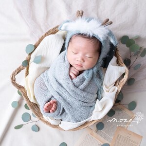 May include: A newborn baby wearing a blue and white knitted deer hat is sleeping in a woven basket. The baby is wrapped in a blue knitted blanket and is surrounded by eucalyptus leaves.