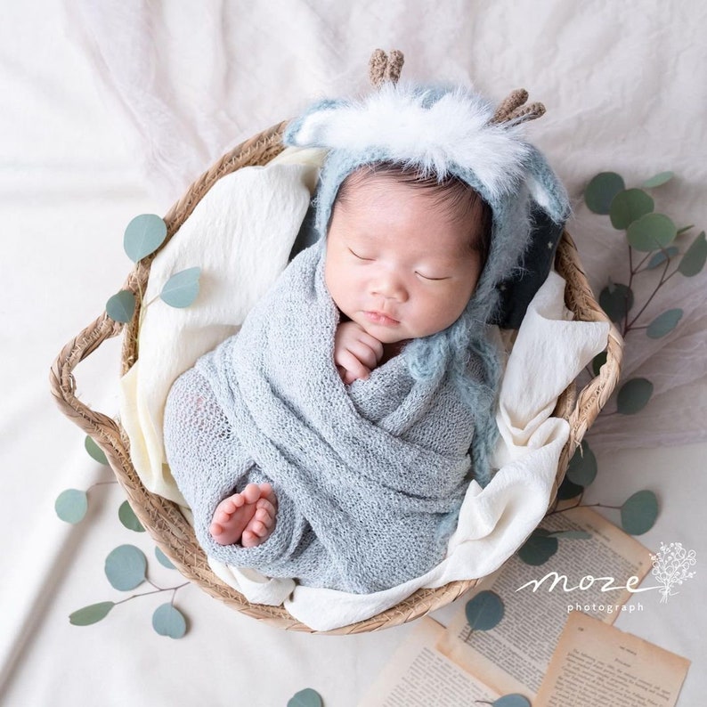 May include: A newborn baby wearing a blue and white knitted deer hat is sleeping in a woven basket. The baby is wrapped in a blue knitted blanket and is surrounded by eucalyptus leaves.