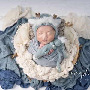 May include: A newborn baby wearing a blue and white deer hat and a blue knitted blanket, holding a blue knitted toy. The baby is lying in a basket surrounded by white and blue blankets.