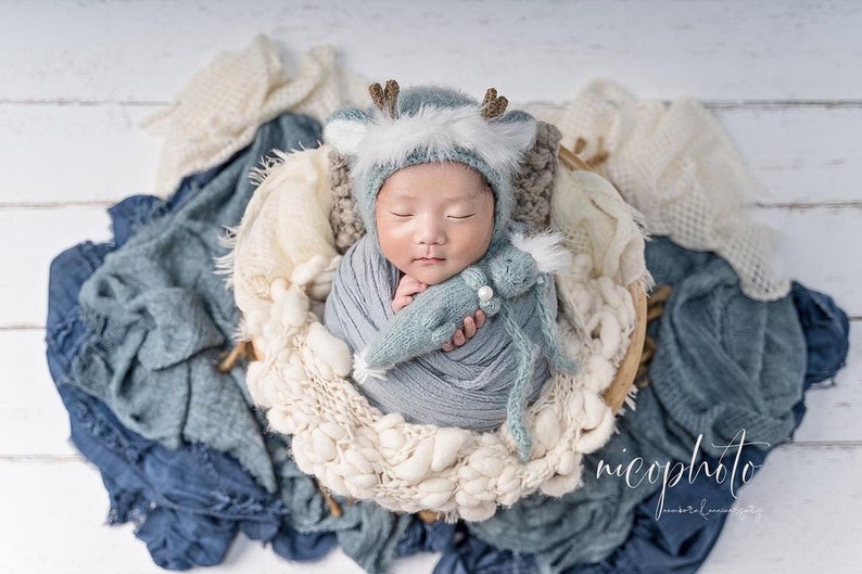 May include: A newborn baby wearing a blue and white deer hat and a blue knitted blanket, holding a blue knitted toy. The baby is lying in a basket surrounded by white and blue blankets.