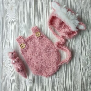 May include: A pink knitted baby outfit with a matching hat. The outfit has two wooden buttons and the hat has a white fur trim.