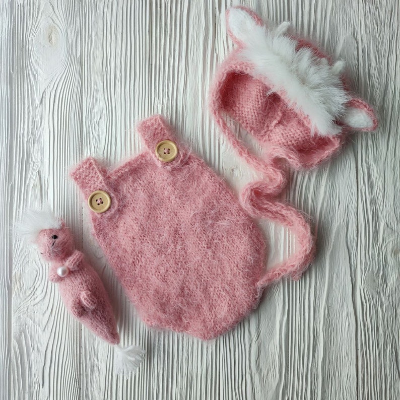 May include: A pink knitted baby outfit with a matching hat. The outfit has two wooden buttons and the hat has a white fur trim.