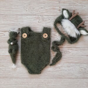 May include: A green knitted baby onesie with two wooden buttons, a matching green knitted hat with white fur trim and antlers, and a small green knitted toy with a pearl.