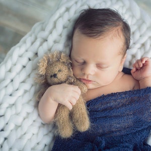 Puede incluir: Un beb&eacute; reci&eacute;n nacido durmiendo pl&aacute;cidamente en una manta de punto blanca, sosteniendo un oso de peluche marr&oacute;n. El beb&eacute; est&aacute; envuelto en una manta azul.